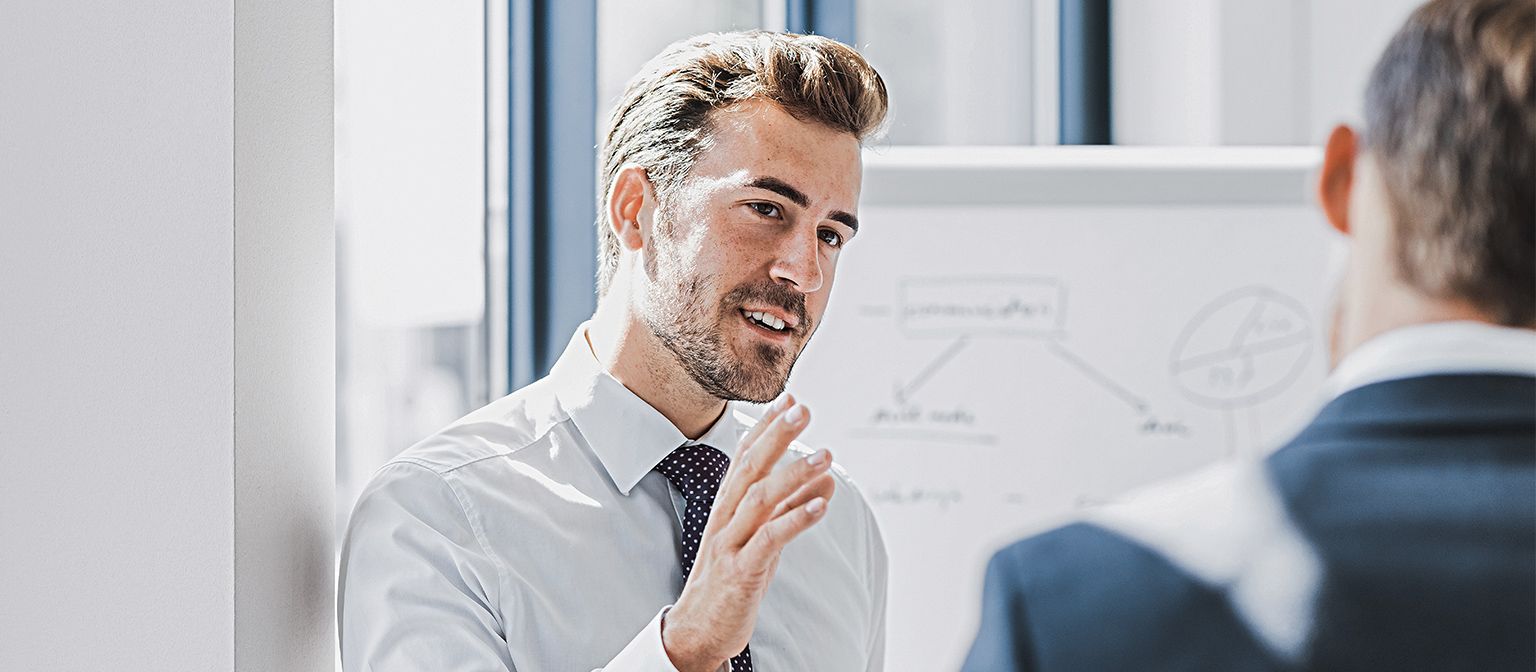 You can see a man in business attire explaining something with a friendly, engaged expression. In front of him stands another person who is clearly listening to him. You can see a whiteboard with a sketch in the background.