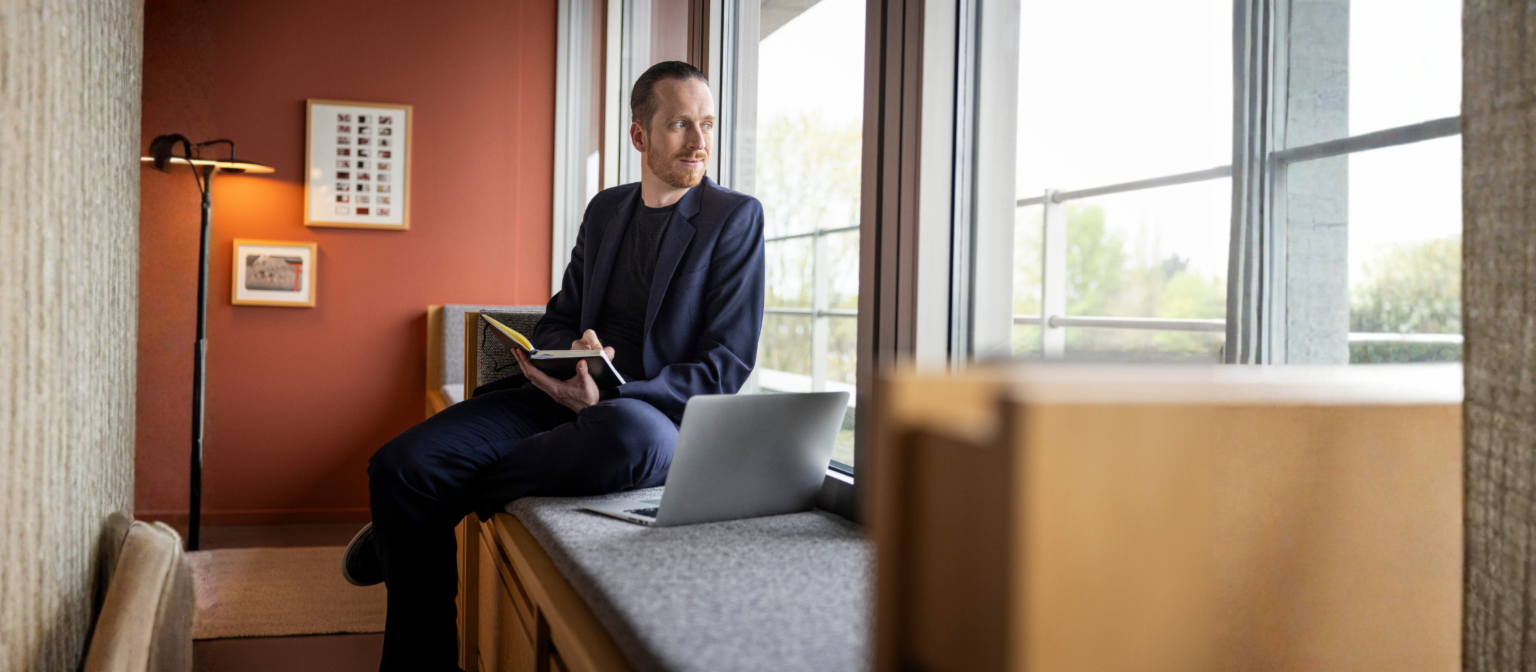 Sebastian Pollmer sits on a windowsill with an open laptop and a notebook in his hand, staring out the window.