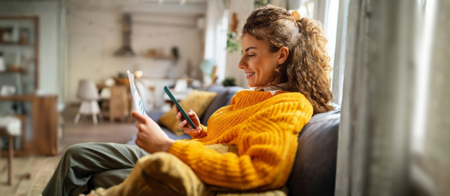 A smiling young woman with curly hair, wearing a cozy yellow sweater, sits on a couch in her living room, looking at her smartphone while holding a paper document.