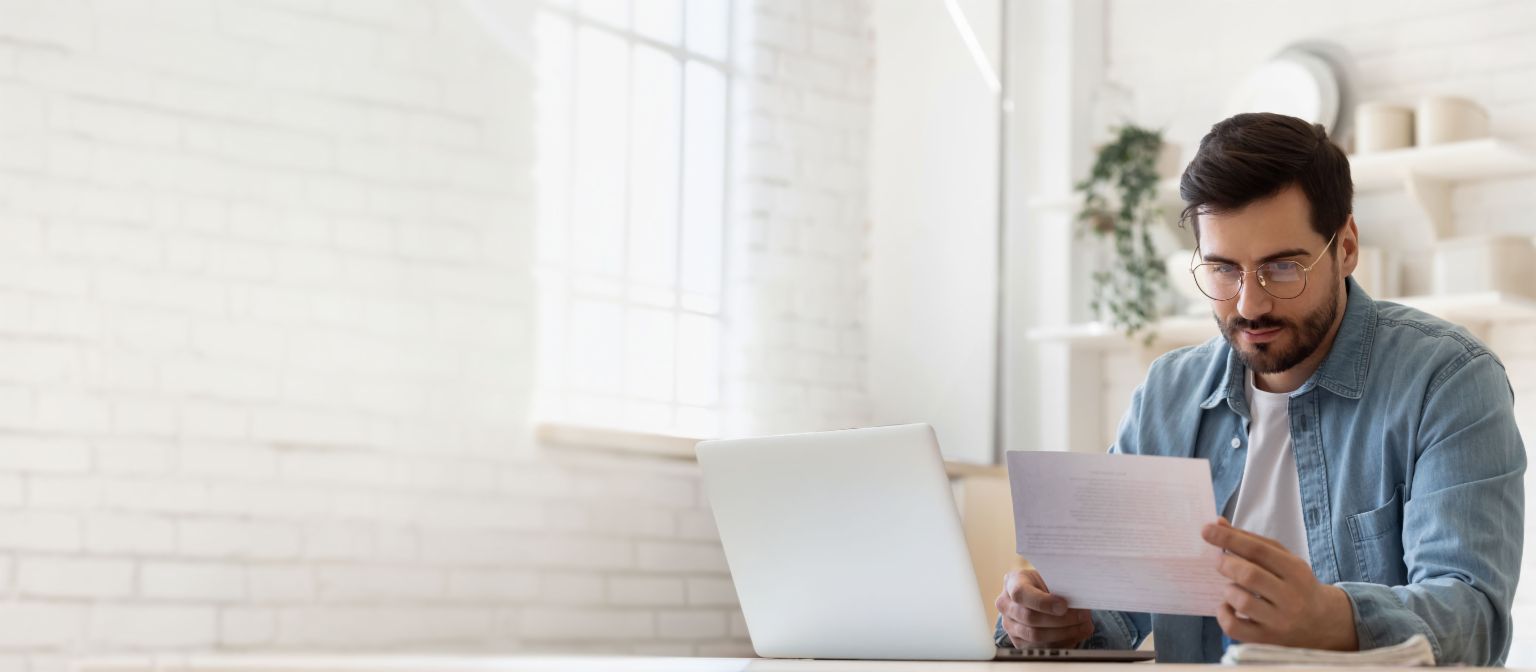 Man wearing glasses working on laptop while reviewing debt settlement document in bright home 