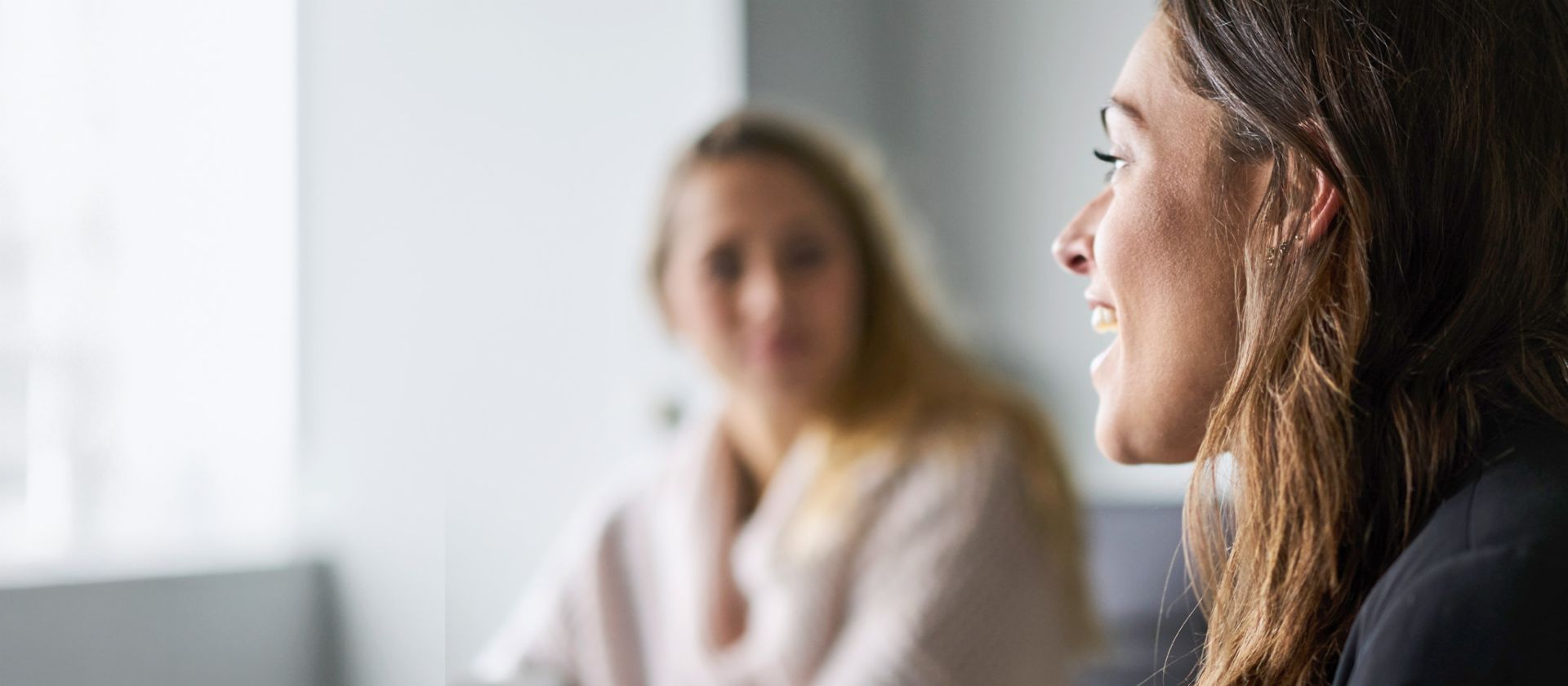 The image features two women in a modern, bright setting, with one woman in the foreground slightly laughing and speaking while looking to the left, and the other in the background turned slightly away, with the background appearing out of focus, showing only their upper bodies.
