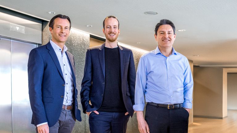 Three male members of the EOS Group’s Board of directors are standing in business attire in front of elevators in a modern setting.