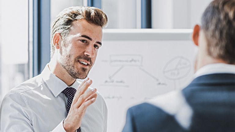 You can see a man in business attire explaining something with a friendly, engaged expression. In front of him stands another person who is clearly listening to him. You can see a whiteboard with a sketch in the background.