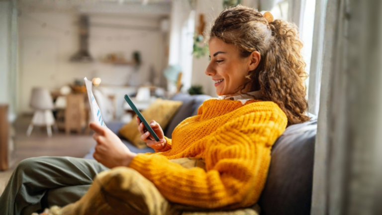 A smiling young woman with curly hair, wearing a cozy yellow sweater, sits on a couch in her living room, looking at her smartphone while holding a paper document.