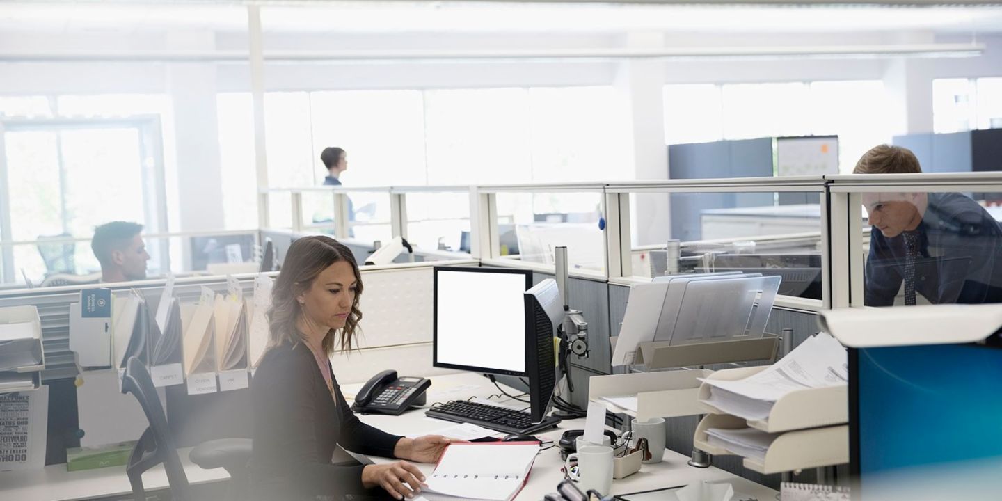 Digitization: Three employees are in an open-plan office; a woman is sitting between papers and her computer