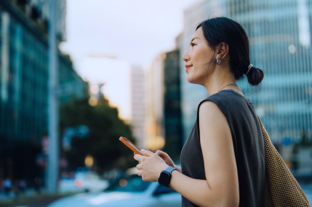 A young woman stands outdoors in an urban environment, holding her smartphone and looking ahead with a smile. Modern buildings and trees are visible in the background.
