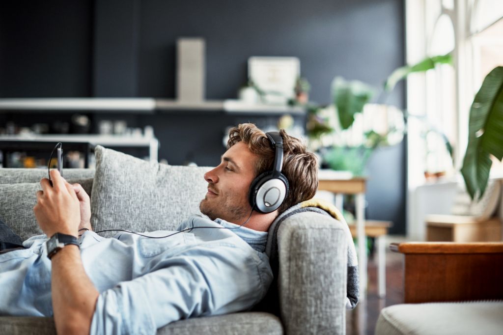 A young man is lying on a couch in his living room, wearing headphones and looking at his smartphone while smiling, enjoying music or a podcast.