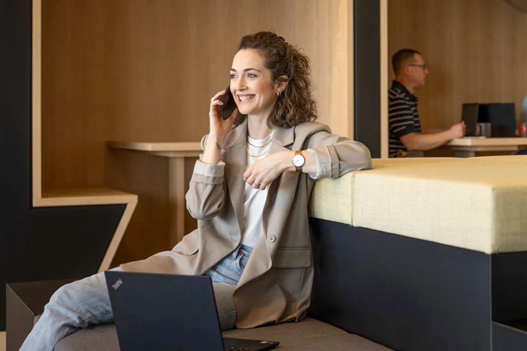 A smiling person sits in a modern office lounge, talking on the phone and working on a laptop. In the background, two other people are working.