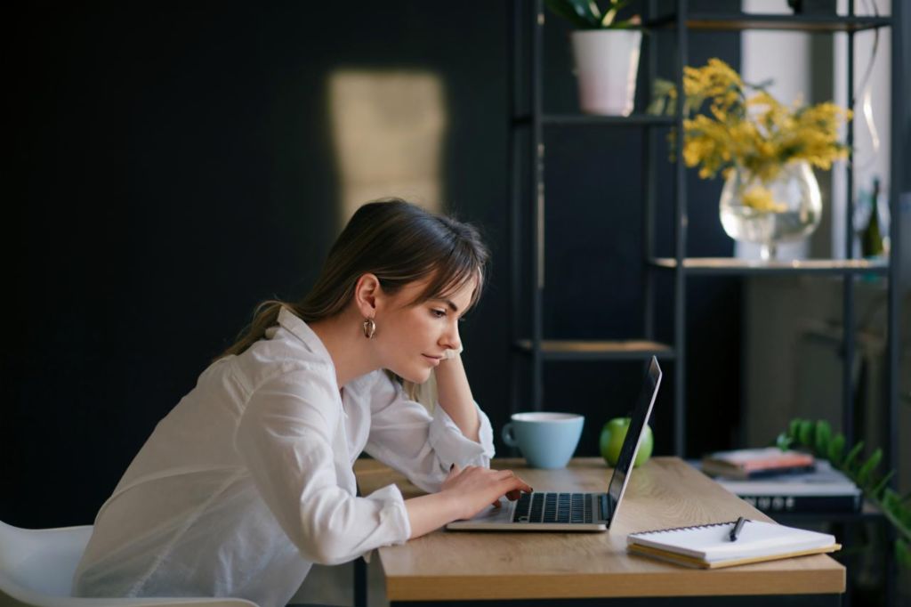 A young woman sits at a desk, working intently on a laptop, with a notebook and a cup beside her. The background is dark, and the focus is on the young woman.