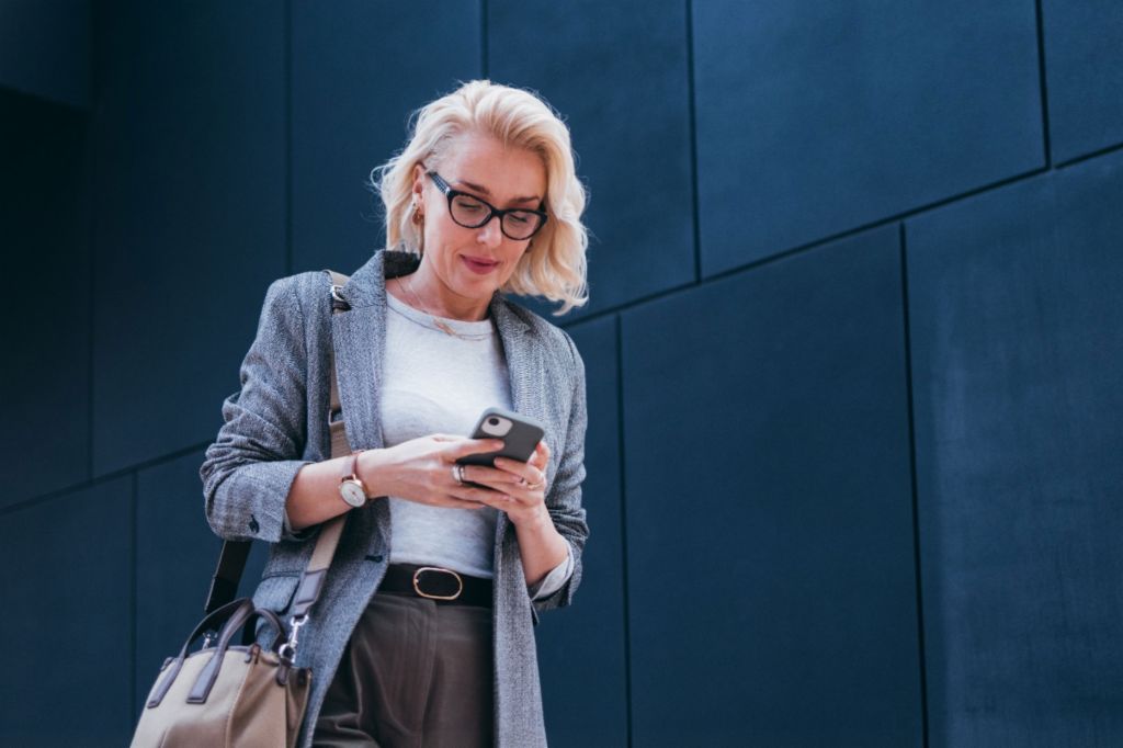 A blonde businesswoman in a grey blazer is standing outside a modern building, looking at her smartphone and smiling.