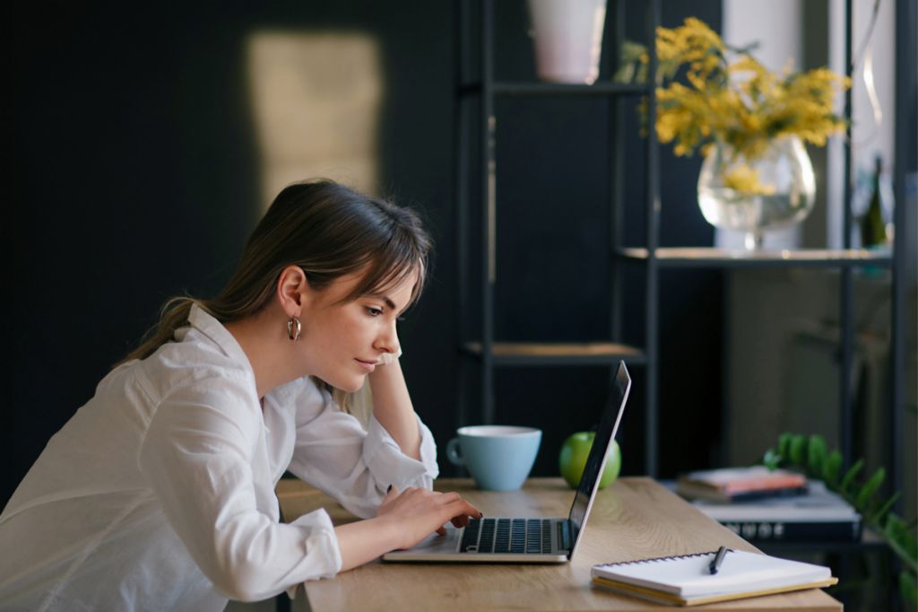 A young woman works intently on a laptop at a desk, with a notebook and a cup beside her.