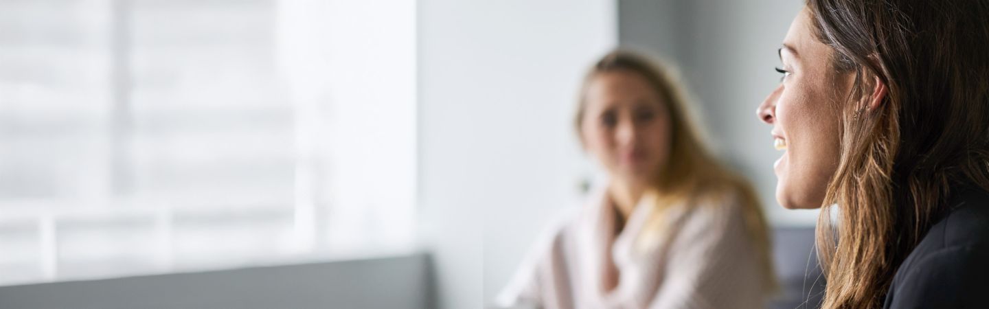 The image features two women in a modern, bright setting, with one woman in the foreground slightly laughing and speaking while looking to the left, and the other in the background turned slightly away, with the background appearing out of focus, showing only their upper bodies.