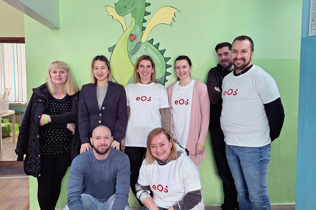 A group of eight people standing in front of a green wall with a painted dragon. Four of them are wearing white t-shirts with an EOS logo printed on the front. 