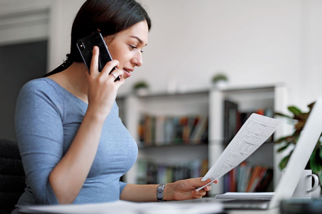 Woman in a blue top on a mobile phone while holding an invoice in her hands